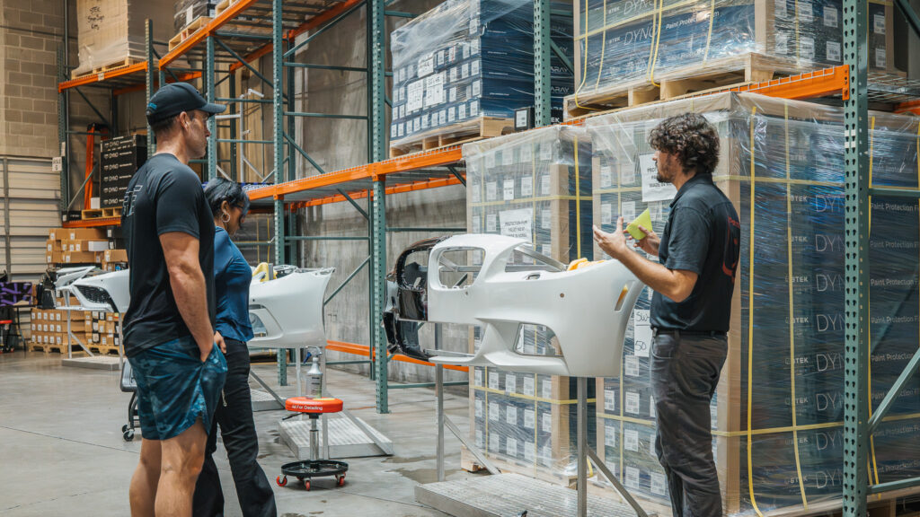 Three workers in a warehouse inspect a white car bumper on a stand among stacked boxes and shelves.