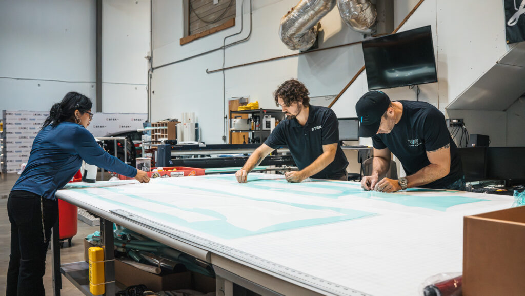 Three people work together on a large panel at a workshop, aligning and smoothing material on a long worktable.