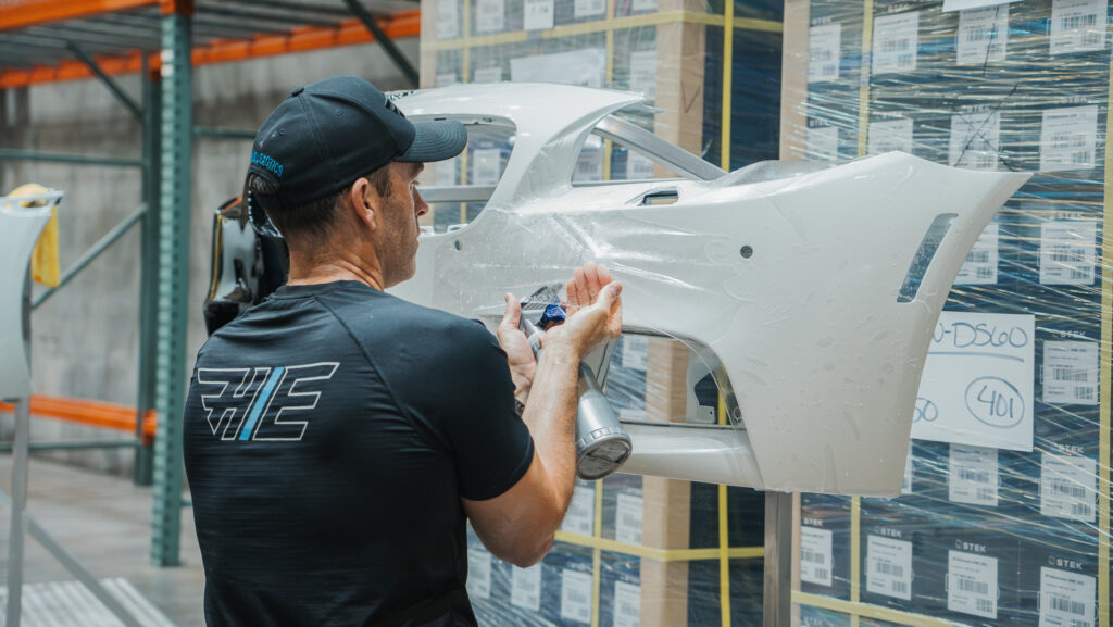 Worker in a cap applying coating to a white car hatch in a warehouse.