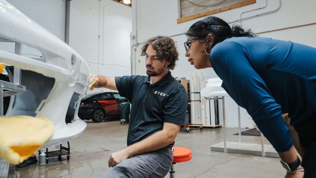Male technician sands a white car bumper while a female colleague in blue observes in a bright auto body shop.
